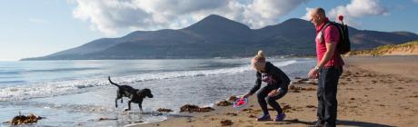 Man and woman with frisbee playing with black dog on beach beside Mourne Mountains