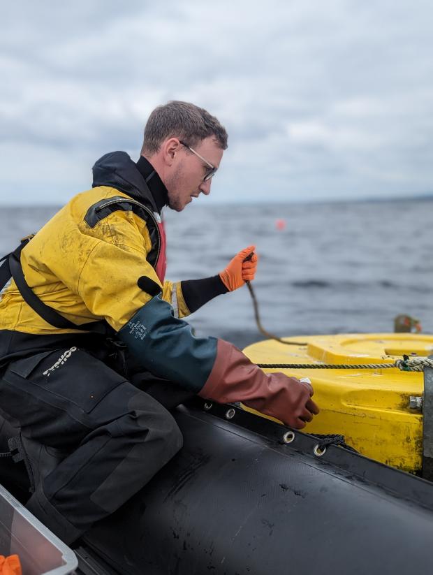 Man in a boat wearing yellow jacket and life vest carrying out maintenance to water monitoring buoy in Lough Neagh
